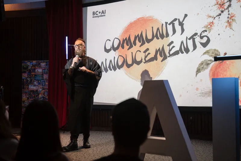 Community Announcements open with Kris on stage beside the peach-framed slide — Wider angle.