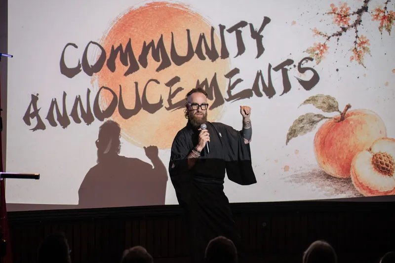Community Announcements open with Kris on stage beside the peach-framed slide — Closer on the mic.