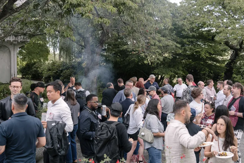 Crowds fill the courtyard for outdoor networking in the late-day light — Screen bright in the background.