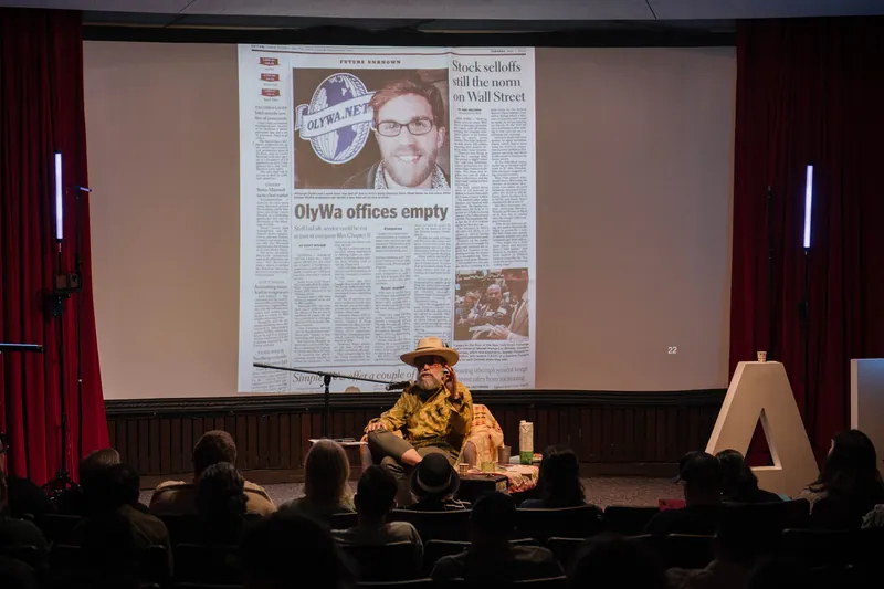 Dave Olson leans into storytelling — clippings, collages, and handwritten notes on screen — Stage-right view.