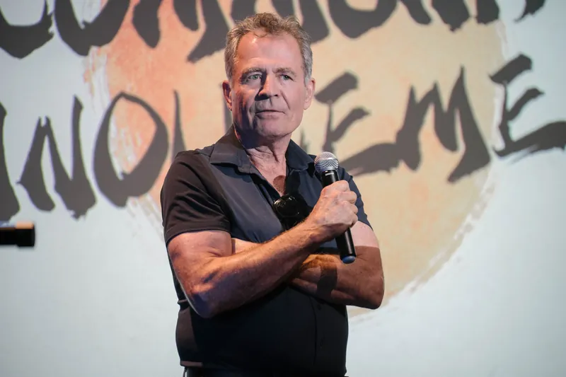 A community announcer in a black tee addresses the room from center stage.