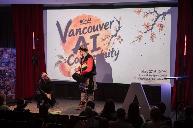 An Indigenous welcome unfolds on stage as the audience settles in — Audience heads in the foreground.