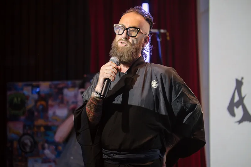 Kris Krüg hosts from the mic in a black wrap jacket, tattoos visible under the stage lights.