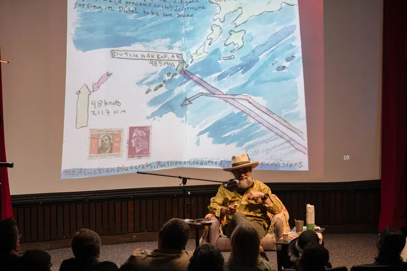 Dave Olson speaks from the armchair, gesturing as slides cycle behind him — Soft focus on the crowd edge.