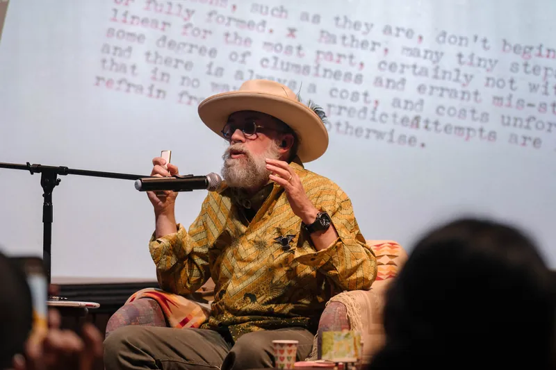 Dave Olson continues from the armchair as denser text and photo slides roll — Audience heads in the foreground.