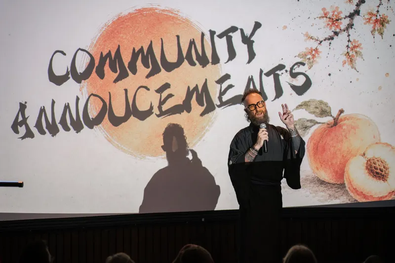 Community Announcements open with Kris on stage beside the peach-framed slide — Soft focus on the crowd edge.