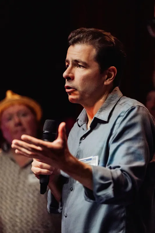 A community presenter gestures with an open hand while holding a microphone.