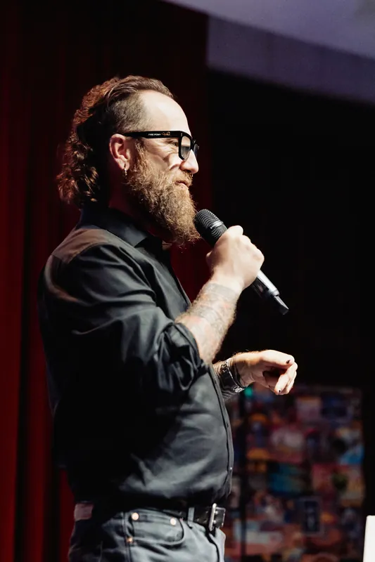 A tight crop captures the host speaking and gesturing onstage.