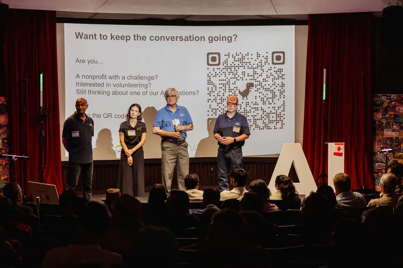 Community presenters stand before a large QR code while the seated audience watches.