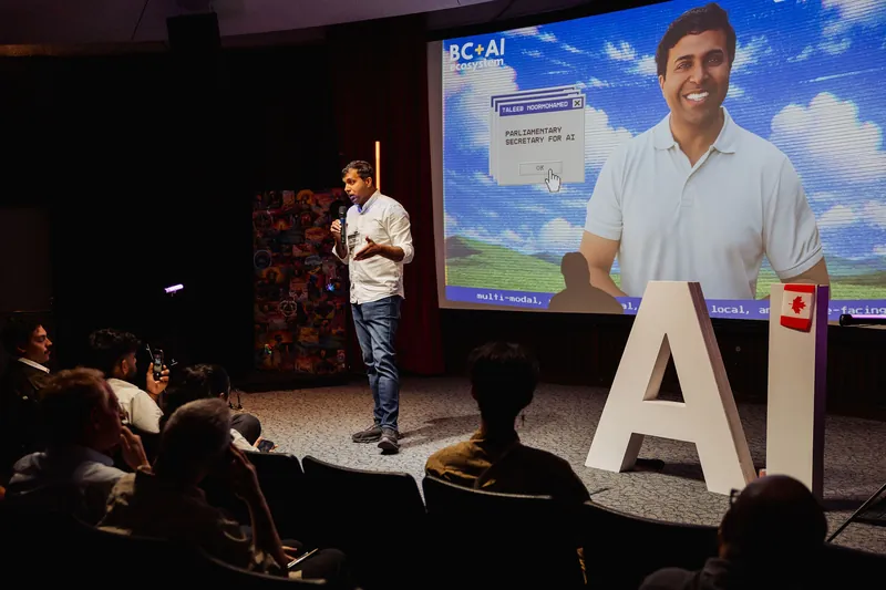 A wide view shows Taleeb Noormohamed speaking to the full seated audience.