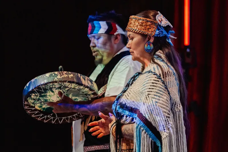Two Indigenous cultural representatives stand together, one holding a decorated drum.