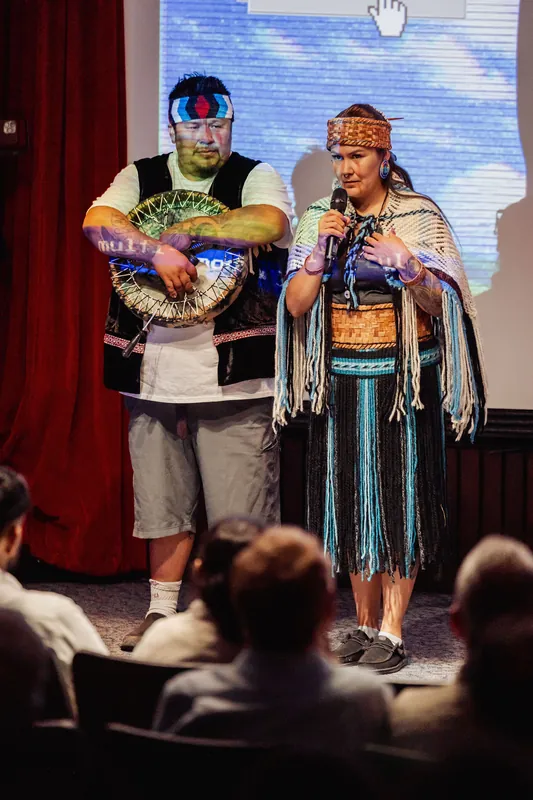 A close view highlights a hand drum and woven ceremonial regalia onstage.