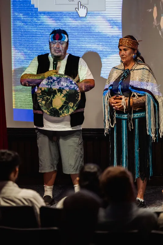 A decorated hand drum and woven ceremonial garments fill a close stage view.