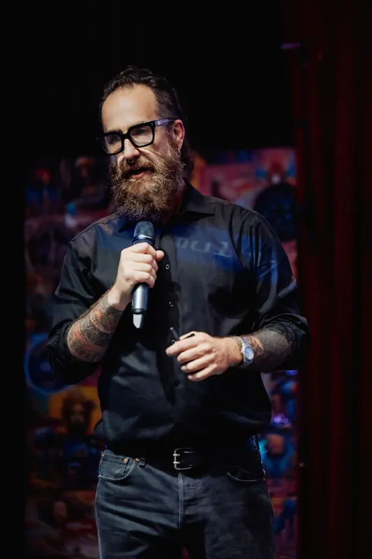 A close detail shows the host holding the microphone between remarks.