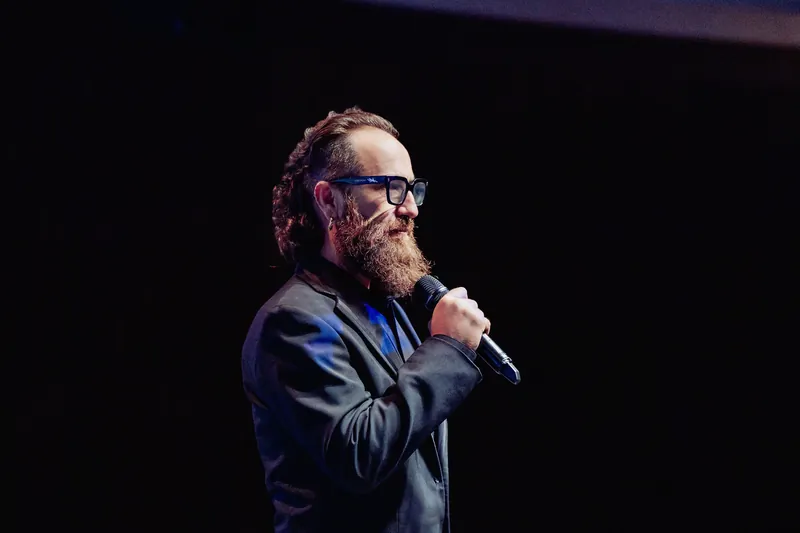 The host speaks in profile against the dark auditorium stage.