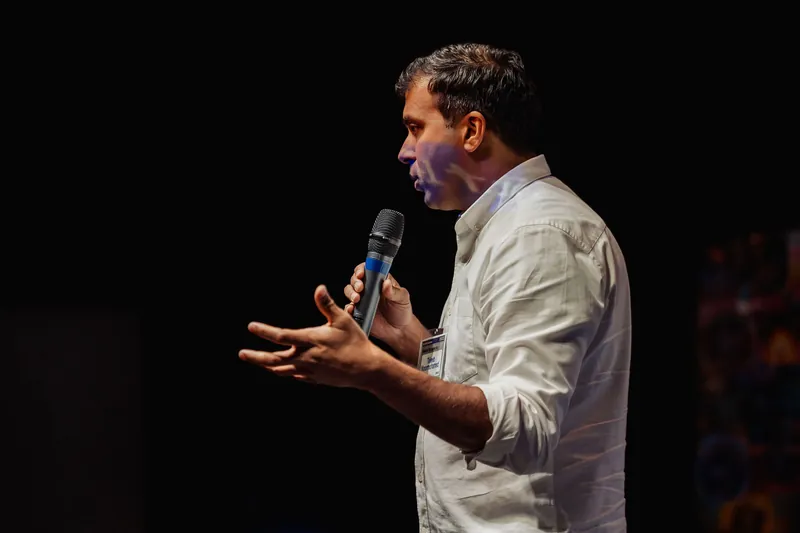 Taleeb Noormohamed addresses the audience with one palm raised.