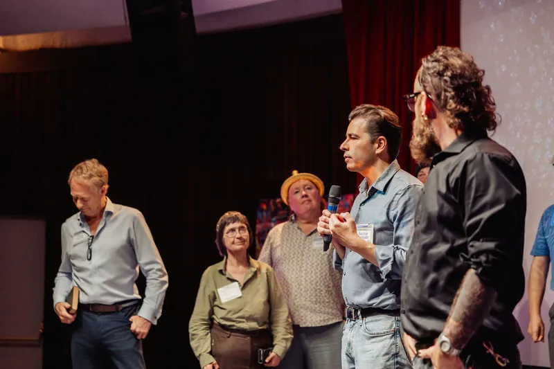 A community presenter gestures with both hands during an onstage announcement.
