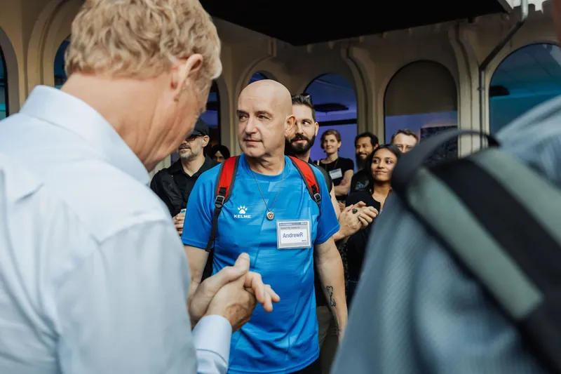 Two attendees talk in the foreground as the indoor crowd gathers behind them.