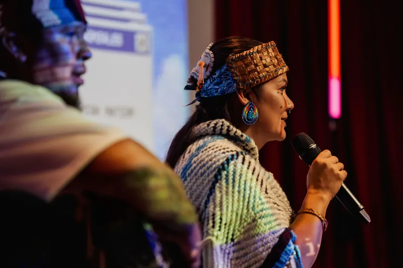 An Indigenous cultural representative addresses the room beside a drum carrier.