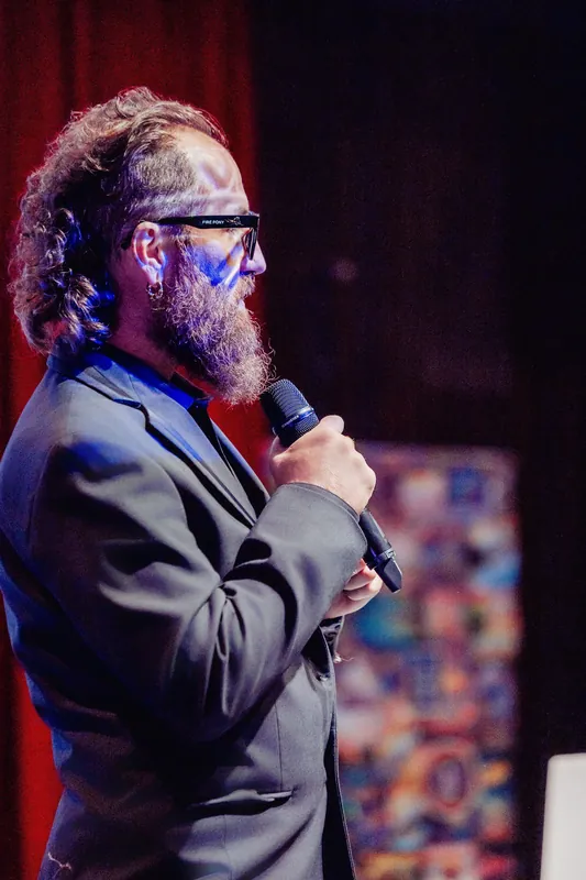 The host speaks into a handheld microphone under colourful stage lights.