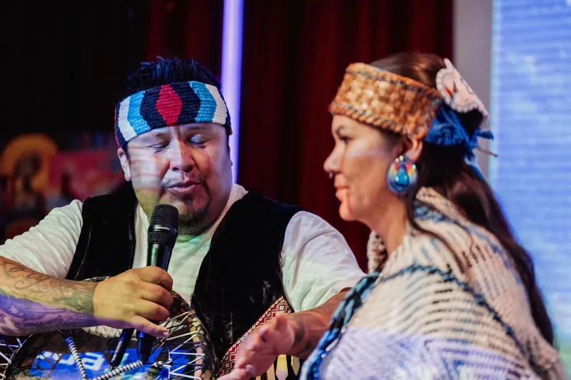 A drum carrier speaks while the second cultural representative listens.