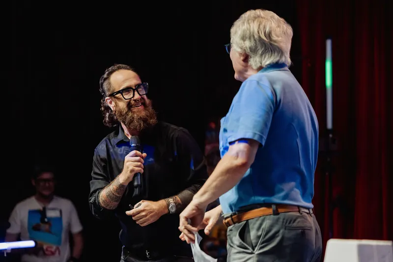 The host and an arriving presenter shake hands onstage.