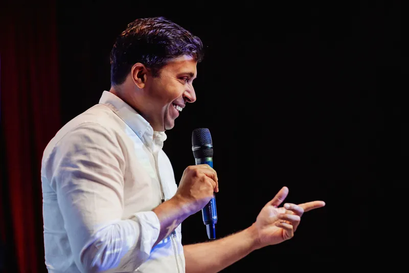 Taleeb Noormohamed speaks into a handheld microphone with one hand extended.