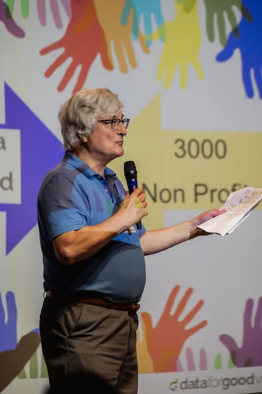 A presenter gestures with printed notes while explaining Data for Good's reach.