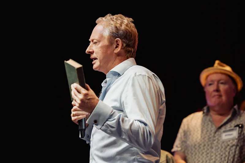 A community presenter holds up a small book while speaking into a microphone.