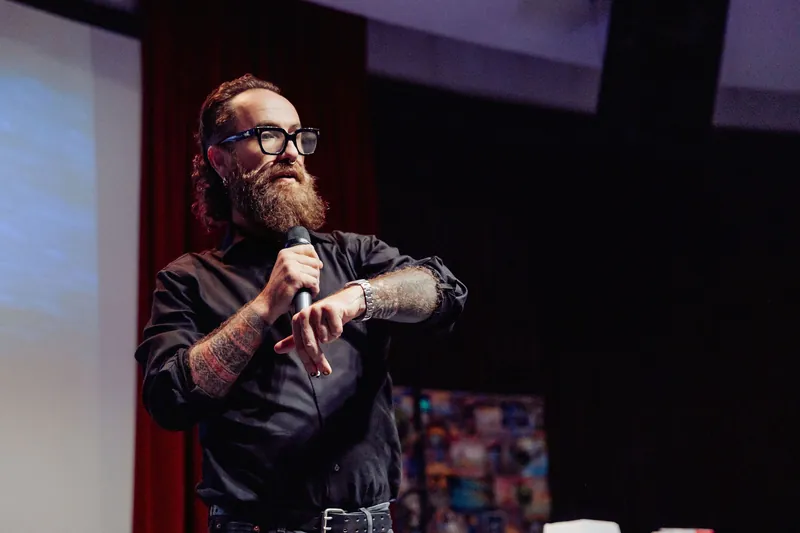 The host addresses the room with one hand resting near the microphone.