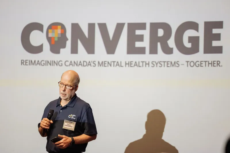 A presenter holds a microphone in front of the CONVERGE mental health slide.