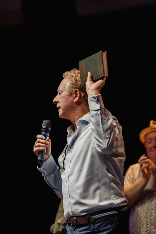 A presenter holds a book high while continuing his community announcement.