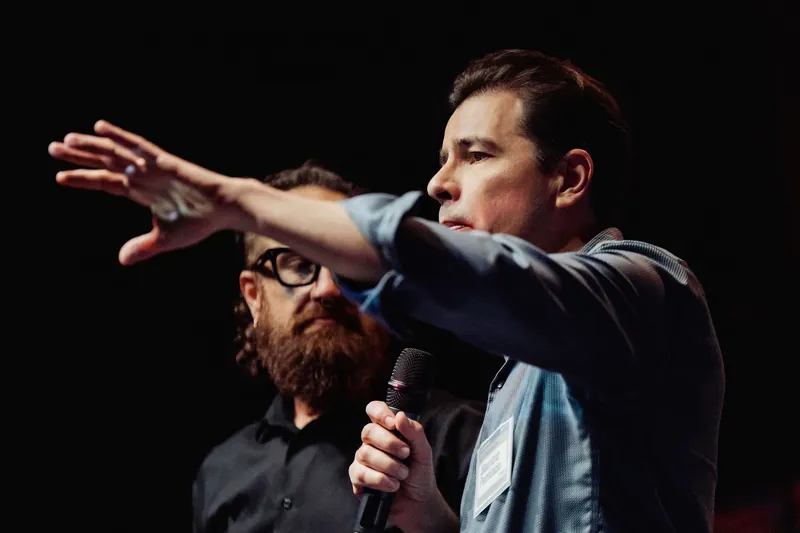 A community presenter extends one arm toward the audience as the host looks on.