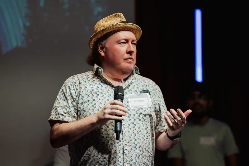 A community presenter in a straw hat speaks into a microphone.