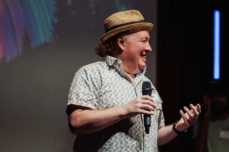 A presenter in a straw hat gestures while addressing the audience.