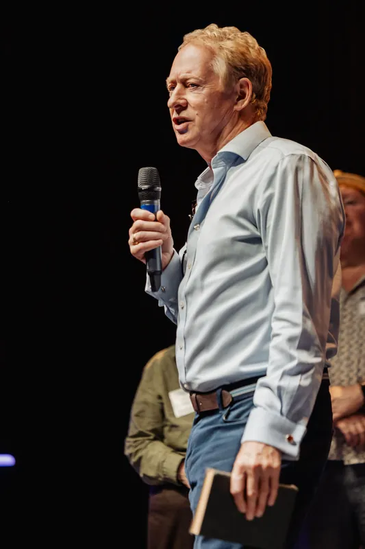 A community presenter speaks into a microphone against the dark stage.
