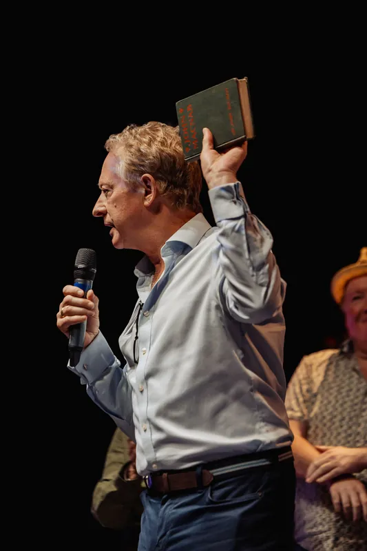 A presenter raises a book and speaks into a handheld microphone.