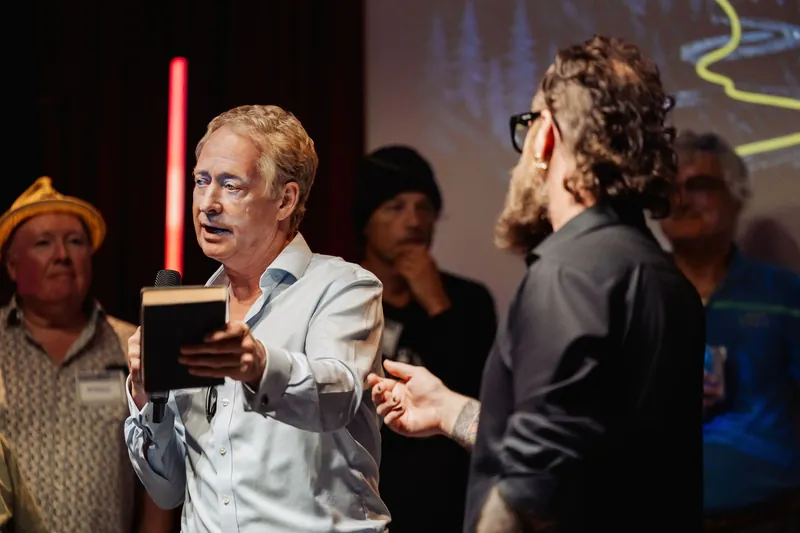 A presenter holds out a small book during a conversation with the host.