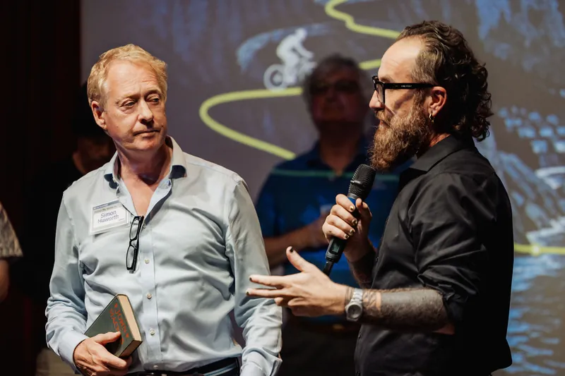 The host speaks into a microphone as the community presenter holds his book.