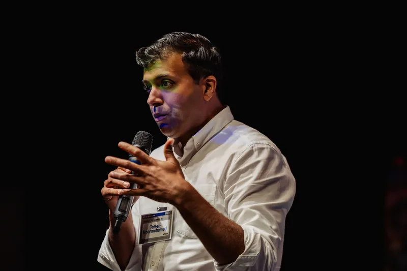 Taleeb Noormohamed holds the microphone close and opens one hand toward the room.