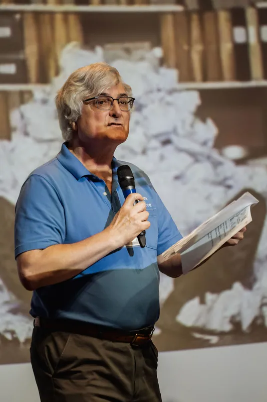 A presenter continues speaking with printed material in hand.