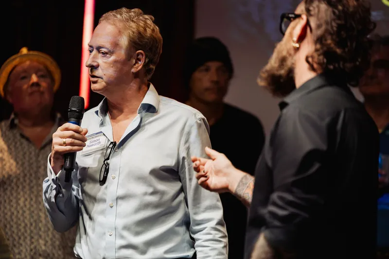 A community presenter speaks beside the host while holding a small book.