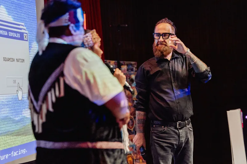 The host gestures while speaking with an Indigenous cultural representative onstage.